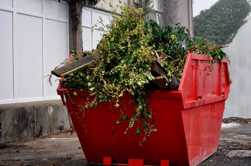 A large red skip bin filled with green foliage, small branches, and garden debris, positioned on a paved outdoor area next to a concrete wall and a white fence with metal framing. The foliage appears to include flowering plants and leafy branches with varying textures, some with small white blossoms. The skip's side panels are made of durable plastic or metal, with visible ridges and a slightly worn finish, indicating it is used for waste disposal. The environment features a mix of urban elements, with the paving surface and adjacent wall suggesting a residential or commercial property in Clapham. The scene is illuminated by natural daylight, highlighting the contrast between the vibrant red container and the green plant material inside. This image illustrates a typical garden clearance or rubbish removal task that a company like Commercial Waste Removal Clapham might service, showcasing the type of waste collected during garden debris removals in the local area.