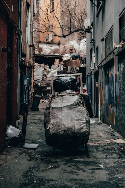 A large outdoor waste collection point featuring multiple open-top wheelie bins and overflowing rubbish. The central bin, made of grey plastic, is filled with mixed paper, cardboard, and assorted pack