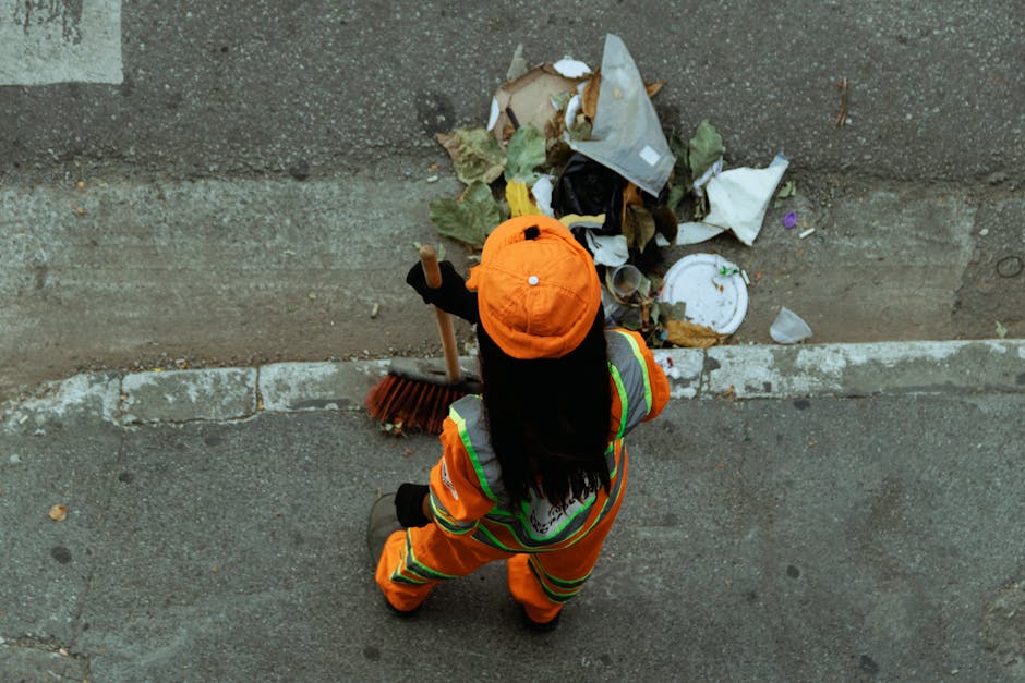 A large green waste disposal bin filled with various construction debris and packaging materials, including cardboard boxes, wooden pallets, and miscellaneous packaging, situated on a city street with